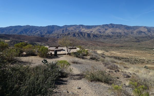 A round stone overlook plaza with a large sundial at a desert rest area, surrounded by shrubs and cactus, with a wide valley and mountains under a clear blue sky