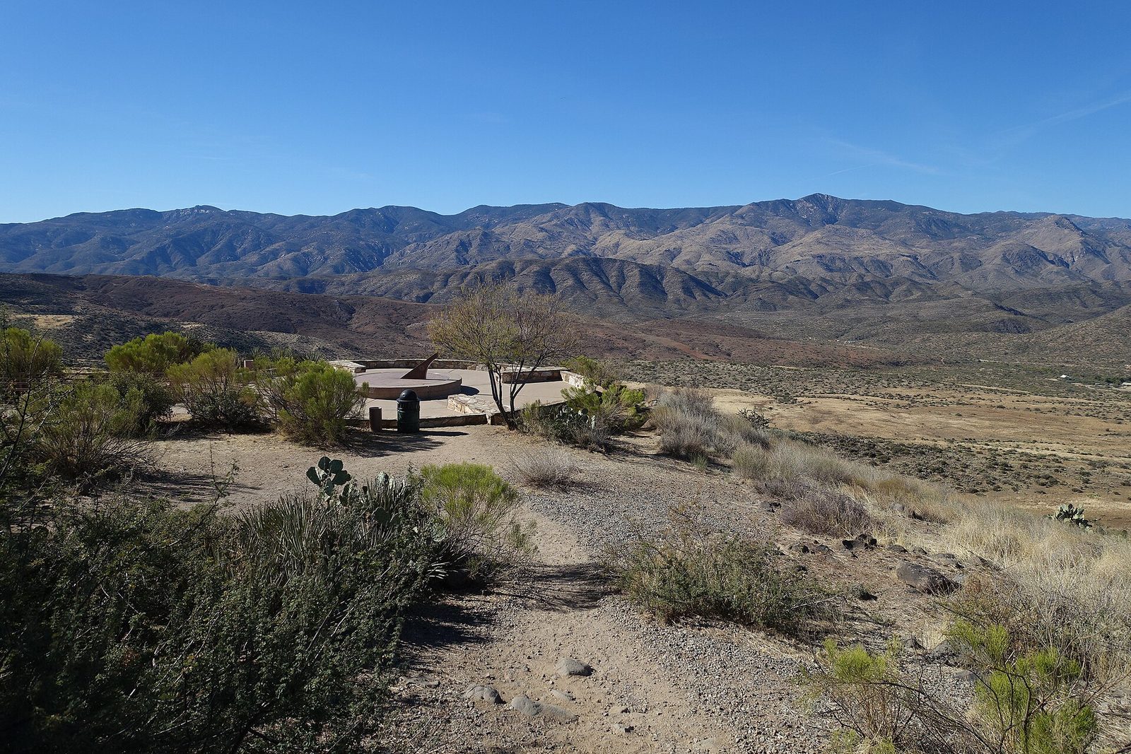 A round stone overlook plaza with a large sundial at a desert rest area, surrounded by shrubs and cactus, with a wide valley and mountains under a clear blue sky