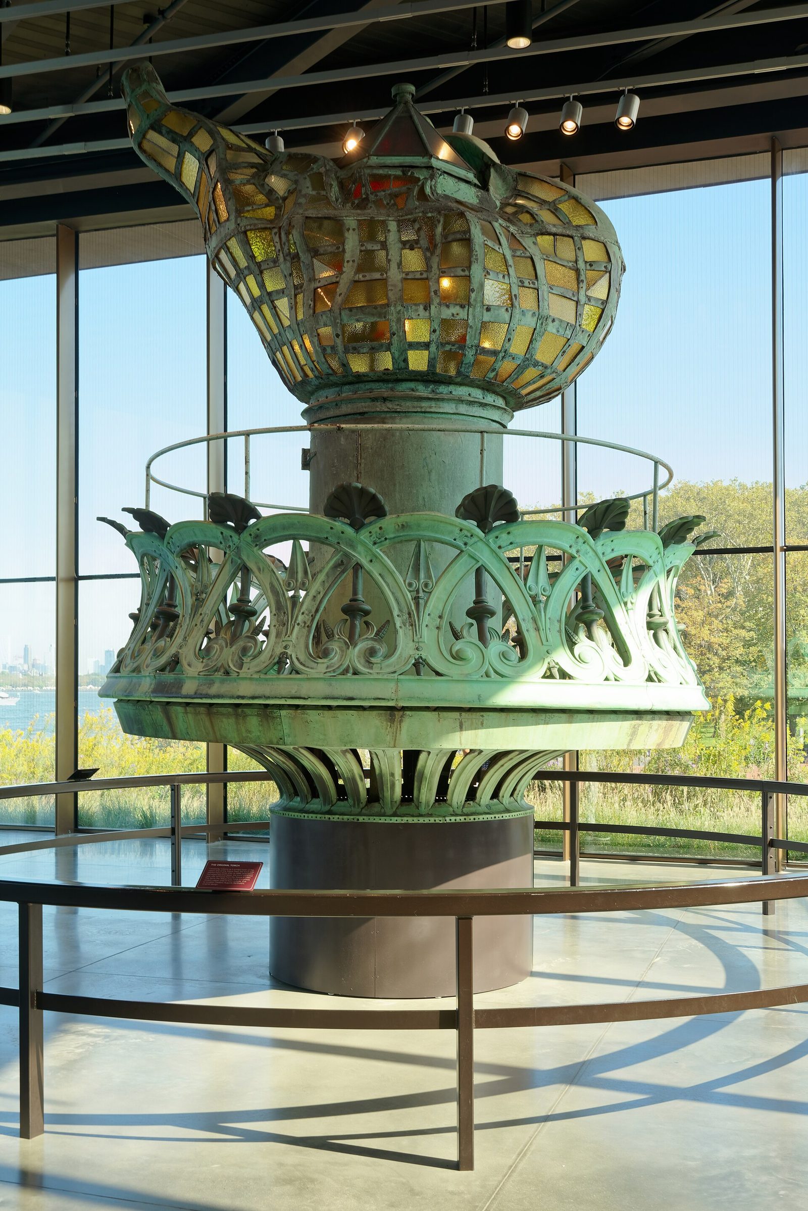 The statue's original copper torch with amber glass panes on display inside a glass-walled museum gallery, with the harbor visible through the windows