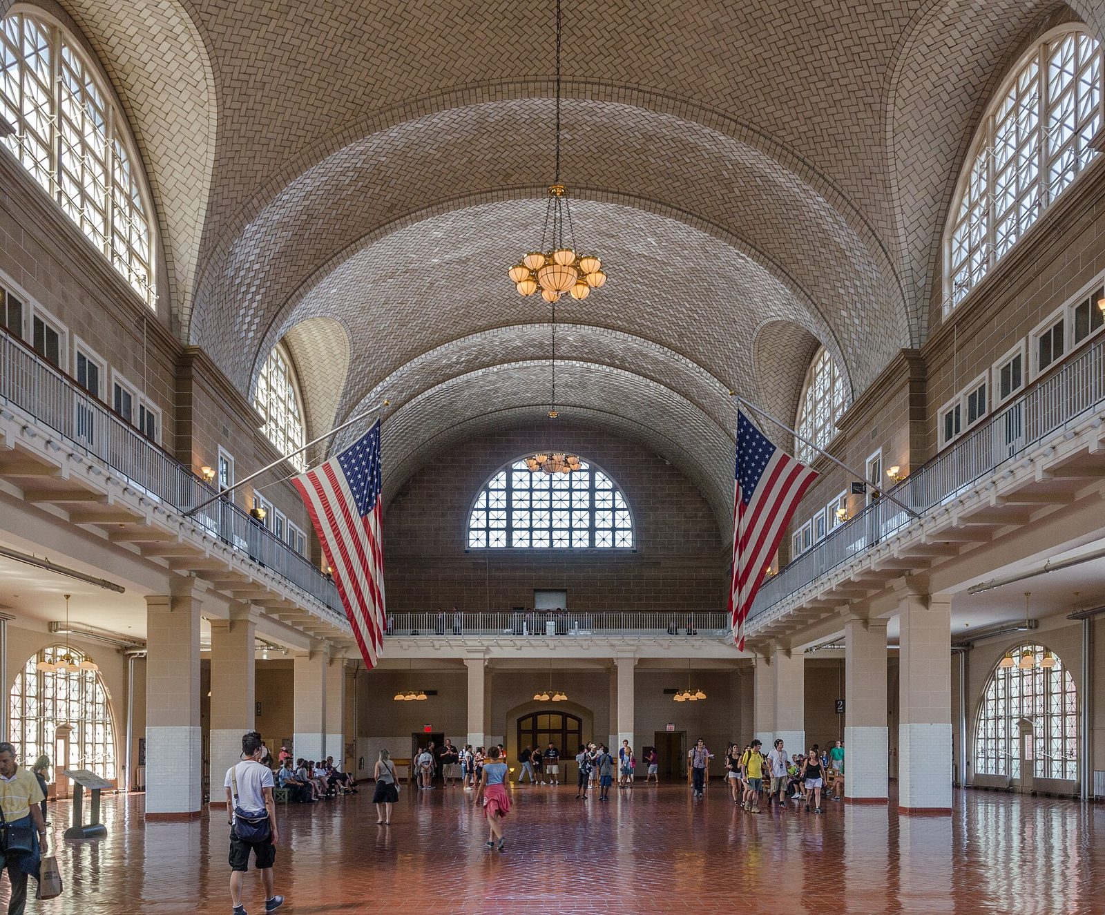The Great Hall at Ellis Island, a long room with a tiled vaulted ceiling, arched windows and two large American flags hanging from the balconies