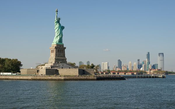 The Statue of Liberty on Liberty Island seen from the ferry on a clear day, with Ellis Island and the Jersey City skyline to the right