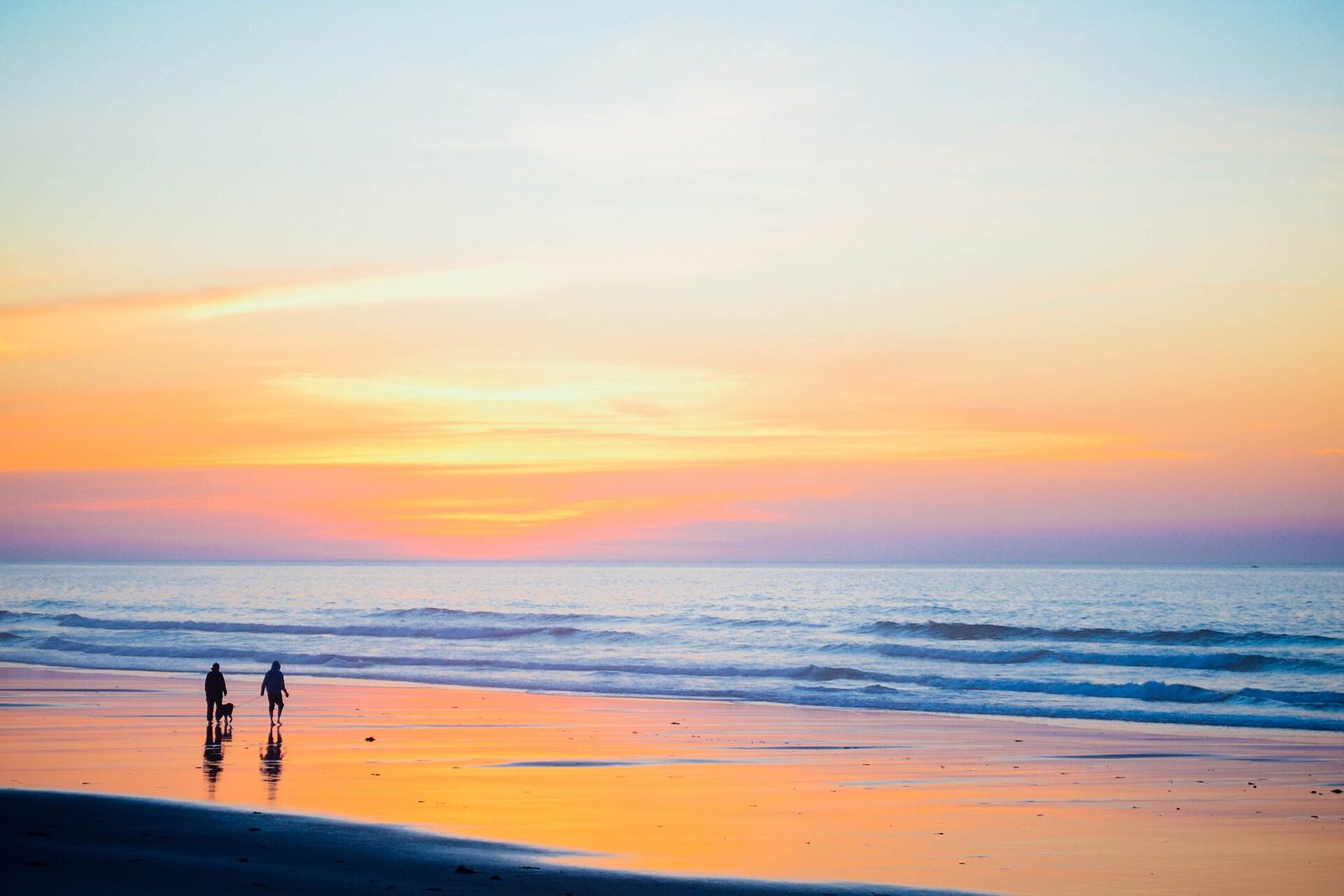 Two people and a dog walking along the wet sand at the edge of the ocean under an orange and pink sunset sky