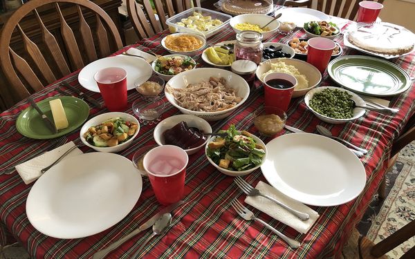 A home dining table with a plaid tablecloth set for Thanksgiving with a bowl of turkey, salads, peas, mashed potatoes, deviled eggs, pies and red cups