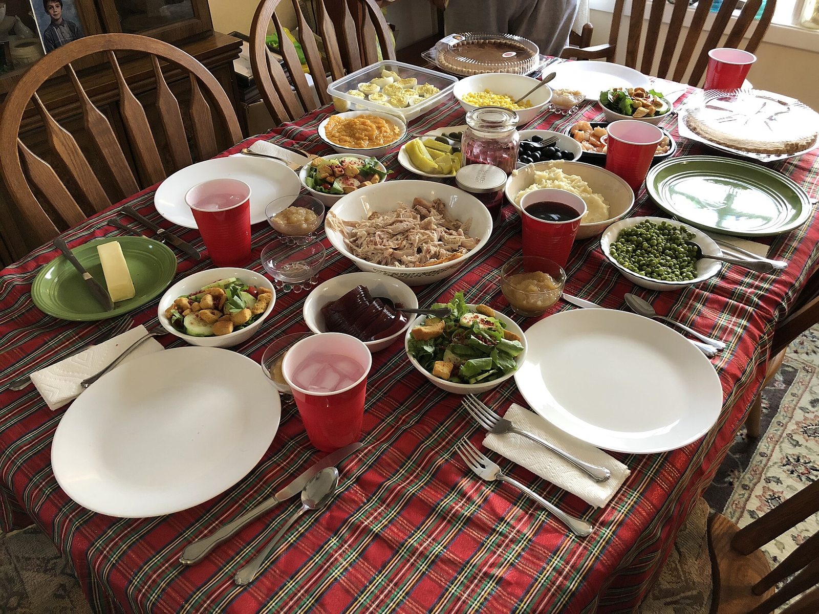 A home dining table with a plaid tablecloth set for Thanksgiving with a bowl of turkey, salads, peas, mashed potatoes, deviled eggs, pies and red cups