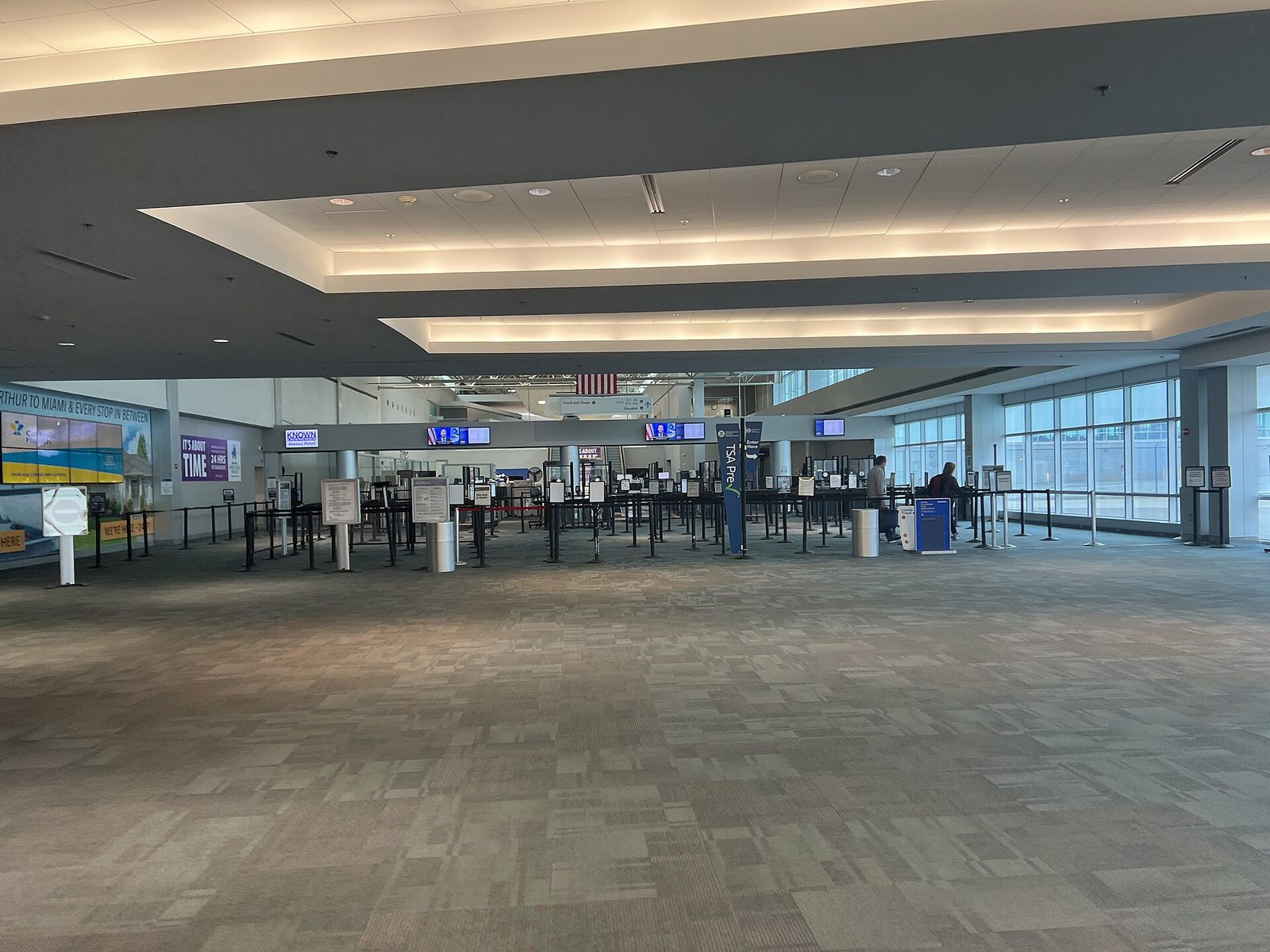 A wide, nearly empty TSA security checkpoint with stanchions, a TSA PreCheck sign and two travelers at the far end