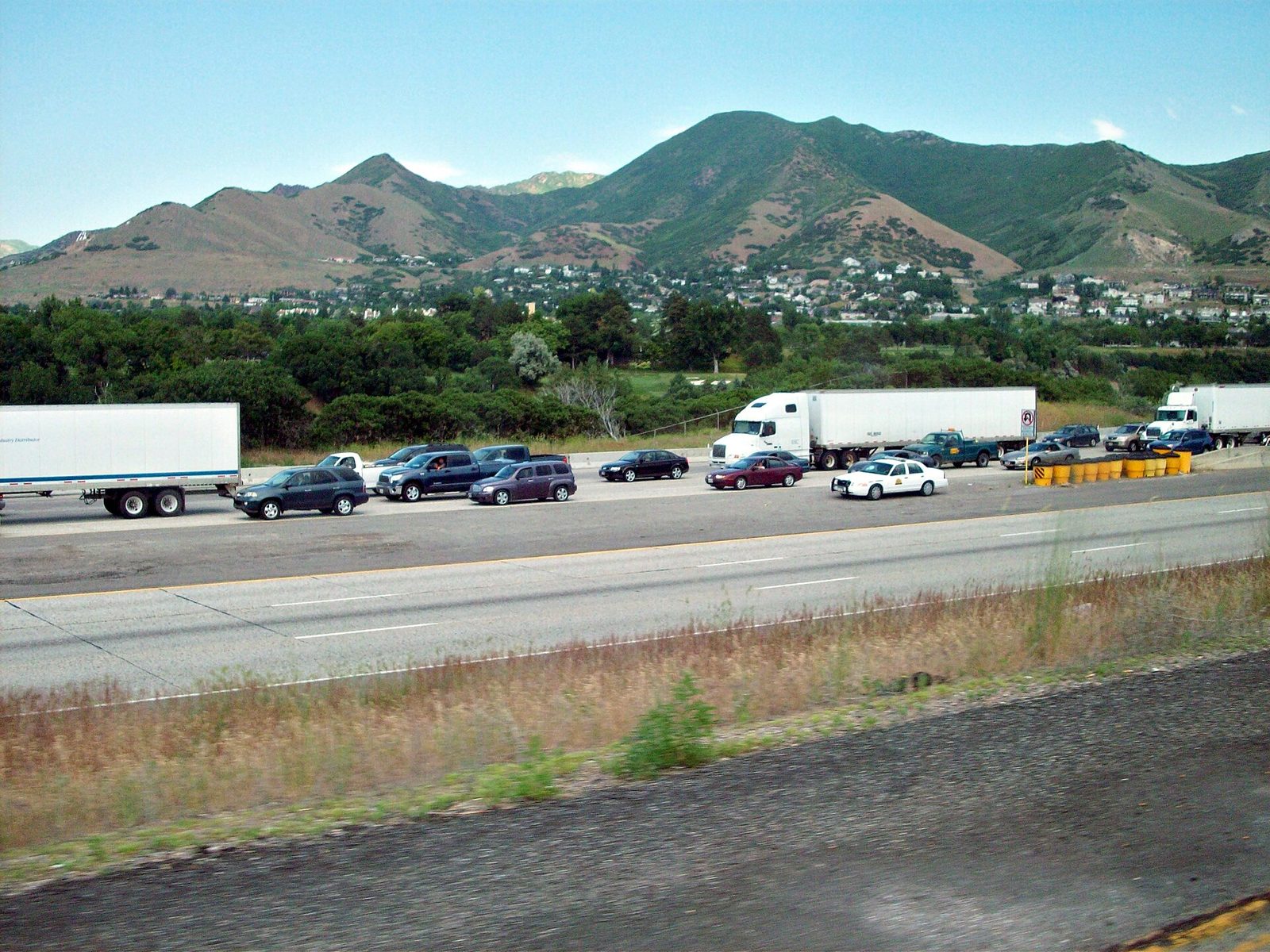 Cars and semi trucks backed up in stop-and-go traffic on a multi-lane interstate with green mountains and houses behind it