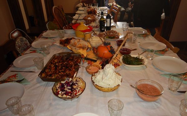 A long holiday table set with white plates, glasses, stuffing, mashed potatoes, cranberry sauce and a small pumpkin centerpiece