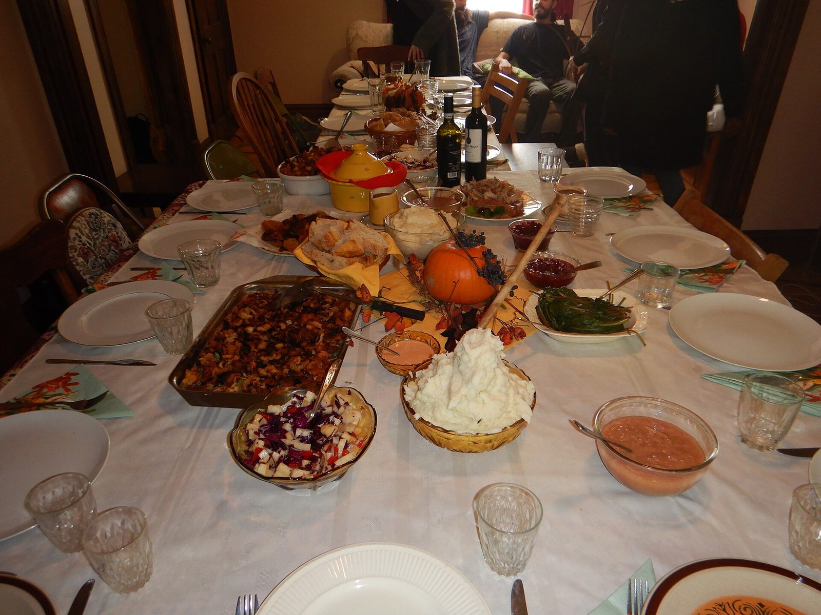 A long holiday table set with white plates, glasses, stuffing, mashed potatoes, cranberry sauce and a small pumpkin centerpiece