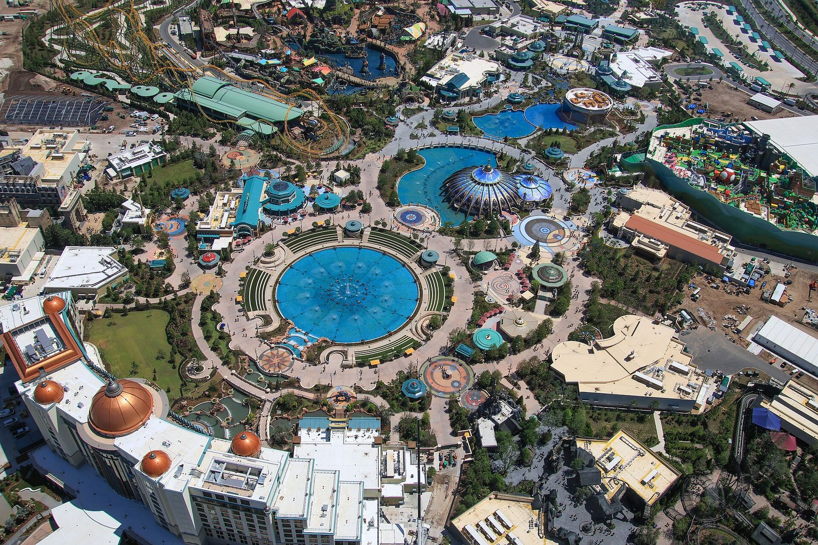 Aerial view of Celestial Park at Universal Epic Universe, with a large round blue fountain at the center, curving paths, a domed pavilion, and roller-coaster track and themed lands around the edges