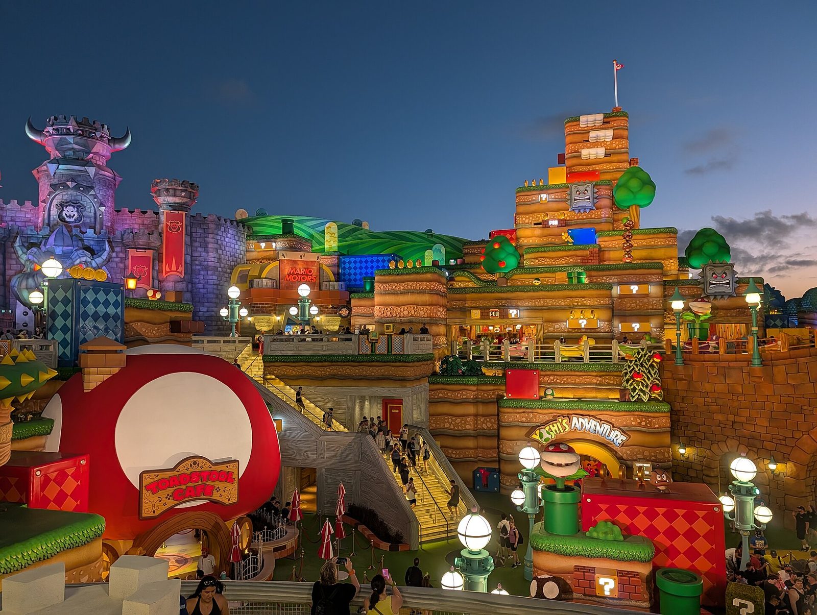Super Nintendo World at Universal Epic Universe at dusk, with Bowser's castle, the Toadstool Cafe mushroom and Yoshi's Adventure lit up and guests on the stairways