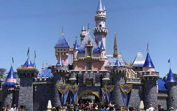 Sleeping Beauty Castle at Disneyland, pink and gray with blue spires under a clear sky, with a crowd of guests in front of the entrance