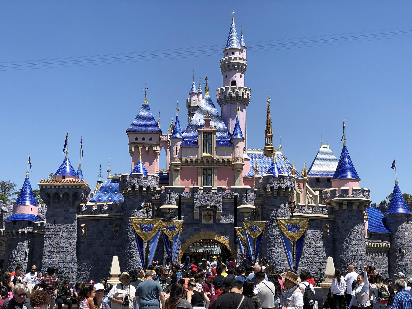 Sleeping Beauty Castle at Disneyland, pink and gray with blue spires under a clear sky, with a crowd of guests in front of the entrance