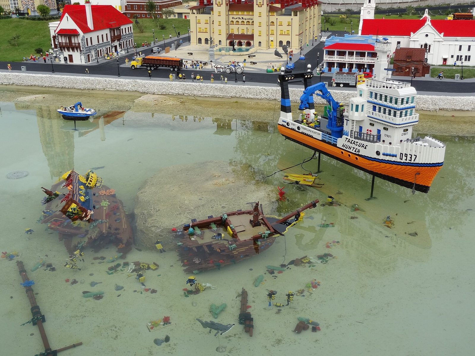 A detailed LEGO brick model of a harbor with an orange and white ship named Treasure Hunter, two sunken wooden shipwrecks in shallow clear water and a miniature town with red roofs behind