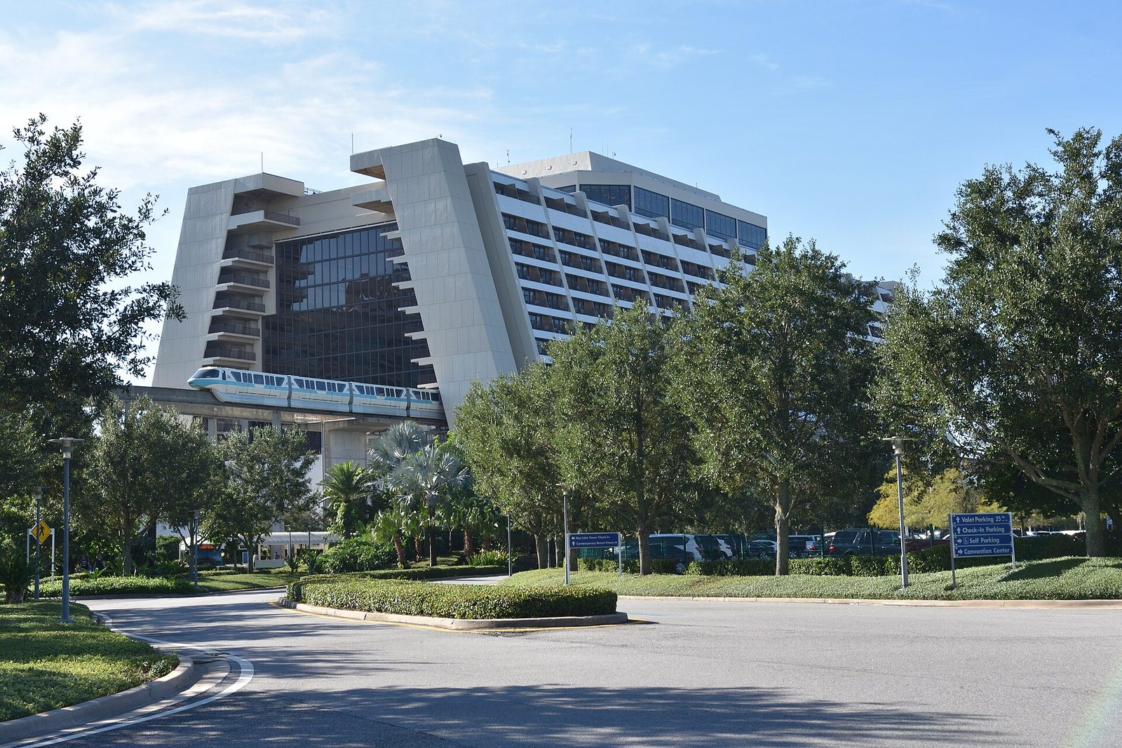 A teal-striped monorail train entering the tall white A-frame Contemporary Resort hotel, with trees and a curving driveway in front