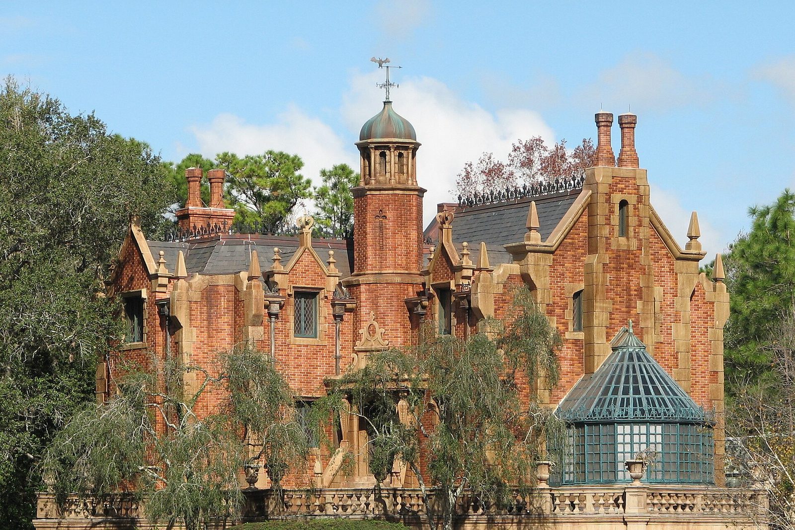 The red-brick Haunted Mansion at Magic Kingdom, with a copper-domed tower, tall chimneys and a glass conservatory, framed by trees under a blue sky