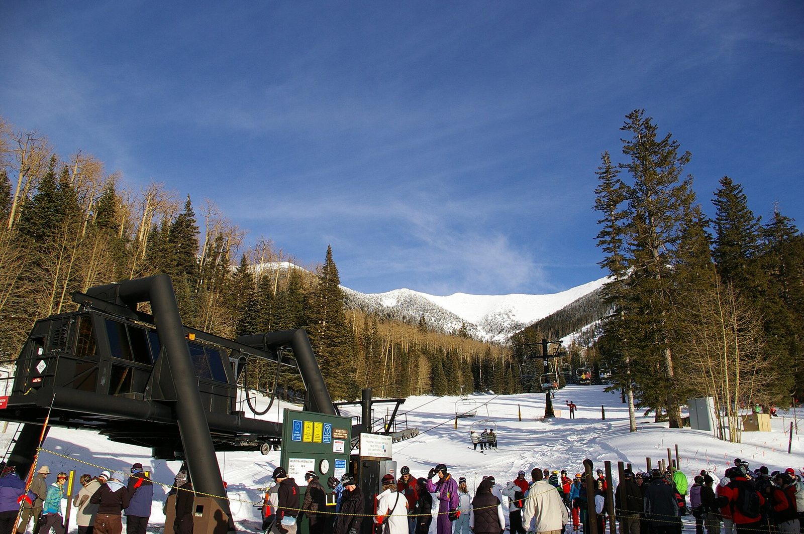 Skiers lining up at the base of a chairlift at Arizona Snowbowl, with snowy peaks and pine trees under a bright blue sky
